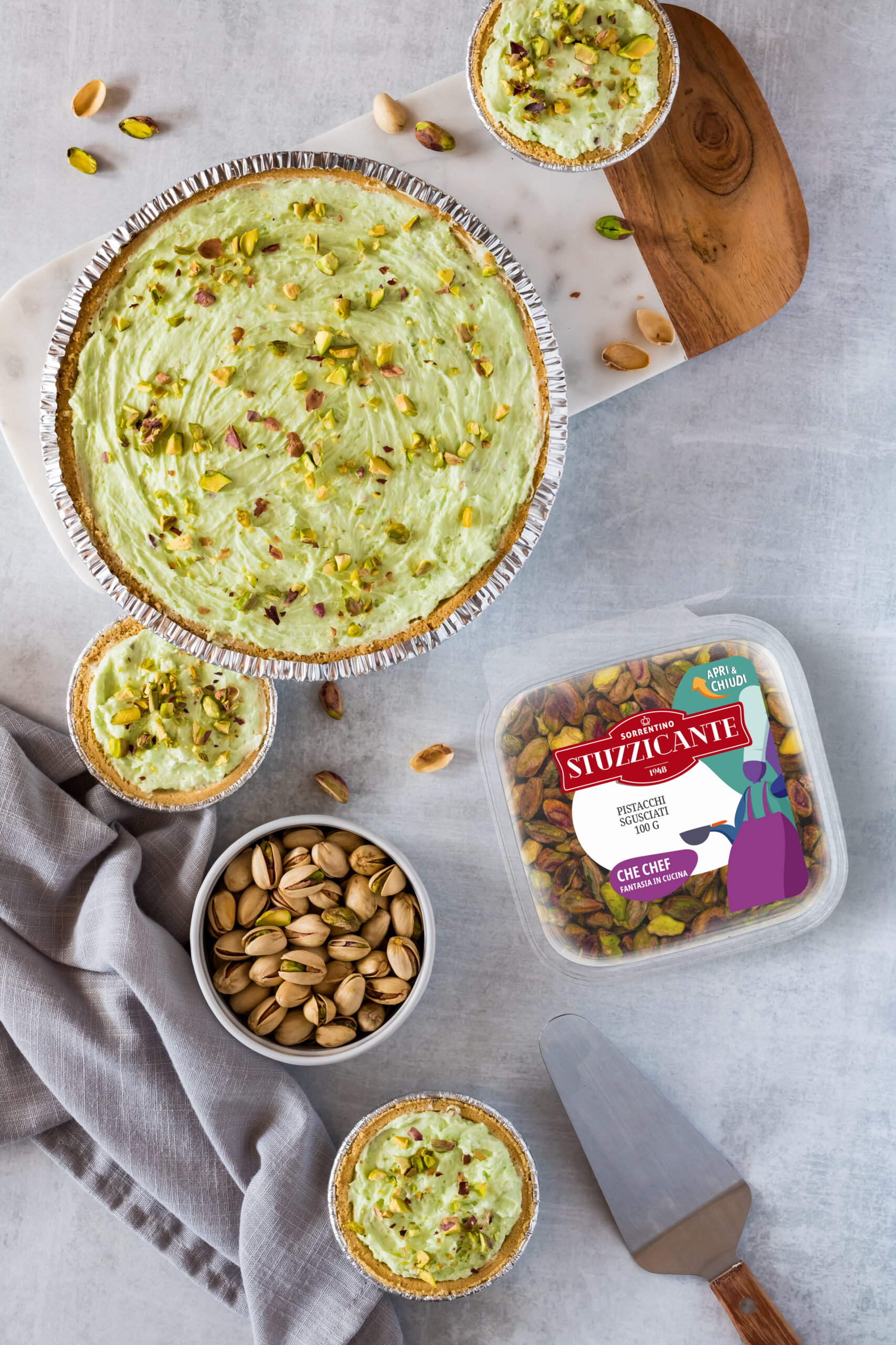 A flat lay view of a variety of pistachio cheesecakes and a bowl of pistachios, with copy space to the right.